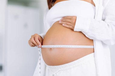 Sıcak bir odada büyüyen göbeğini ölçen hamile bir kadının yakın çekimi. Bir çocuk bekleyen kadın, karnının ölçüsünü şeritle ölçer. Samimiyet kavramı, çocuk doğurmaya hazırlık..