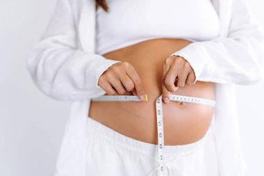 Sıcak bir odada büyüyen göbeğini ölçen hamile bir kadının yakın çekimi. Bir çocuk bekleyen kadın, karnının ölçüsünü şeritle ölçer. Samimiyet kavramı, çocuk doğurmaya hazırlık..