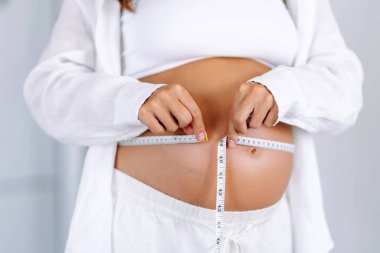 Sıcak bir odada büyüyen göbeğini ölçen hamile bir kadının yakın çekimi. Bir çocuk bekleyen kadın, karnının ölçüsünü şeritle ölçer. Samimiyet kavramı, çocuk doğurmaya hazırlık..