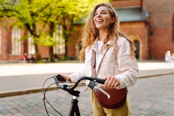 Güneşli bir caddede bisikletli neşeli genç bir kadın duruyor. Güzel bir kadın gün batımını sever, bisiklet sürer. Turizm, eğlence konsepti. Yaşam biçimi.