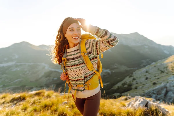 Sırt çantalı kadın yürüyüşçü, gün batımında dağ manzarasına hayran kalarak dağın tepesinde duruyor. Gezgin özgürlüğü hissediyor, dağlarda güneş ışınlarının tadını çıkarıyor. Macera konsepti, doğa.