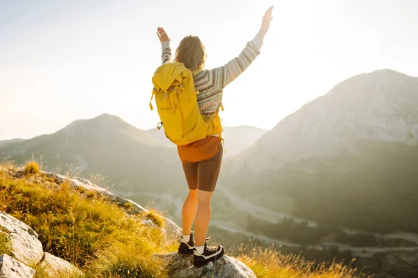 Sırt çantalı kadın yürüyüşçü, gün batımında dağ manzarasına hayran kalarak dağın tepesinde duruyor. Gezgin özgürlüğü hissediyor, dağlarda güneş ışınlarının tadını çıkarıyor. Macera konsepti, doğa.