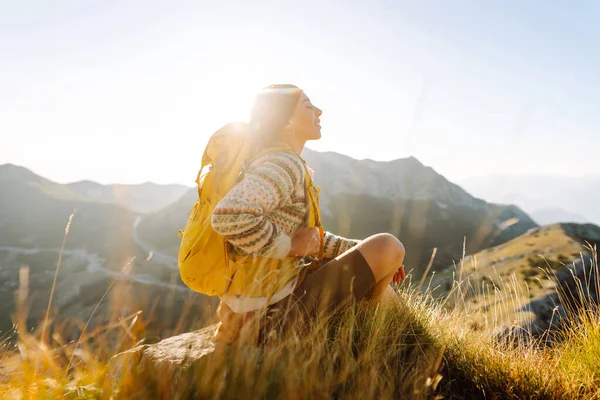 Sırt çantalı kadın yürüyüşçü, gün batımında dağ manzarasına hayran kalarak dağın tepesinde duruyor. Gezgin özgürlüğü hissediyor, dağlarda güneş ışınlarının tadını çıkarıyor. Macera konsepti, doğa.