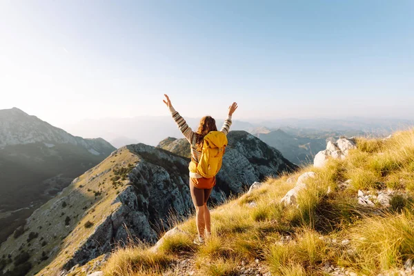 Sırt çantalı kadın yürüyüşçü, gün batımında dağ manzarasına hayran kalarak dağın tepesinde duruyor. Gezgin özgürlüğü hissediyor, dağlarda güneş ışınlarının tadını çıkarıyor. Macera konsepti, doğa.