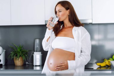 Güzel bir kadın, elinde bir bardak suyla modern mutfakta çocuk bekliyor. Hamile kadın su içiyor ve rahat bir odada dinleniyor. Sağlık ve refah kavramı.