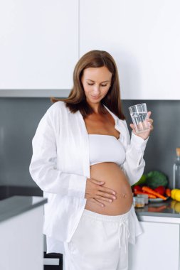 Güzel bir kadın, elinde bir bardak suyla modern mutfakta çocuk bekliyor. Hamile kadın su içiyor ve rahat bir odada dinleniyor. Sağlık ve refah kavramı.