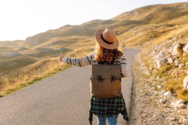 Şapkalı bir yürüyüşçü yol kenarında durup, güneşli bir günde dağ manzarasının tadını çıkarıyor. Genç bir kadın otostop çekiyor, dağlarda eğleniyor. Açık hava aktiviteleri konsepti, doğa.