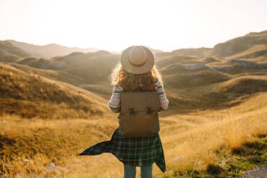 Sırt çantası ve şapkası olan kadın gezgin gün batımında dağ manzarasının tadını çıkarıyor. Güneşli bir günde kayalık bir yolda yürüyen bir turist kendini özgür hisseder. Seyahat etme fikri, özgürlük. Etkin yaşam biçimi.