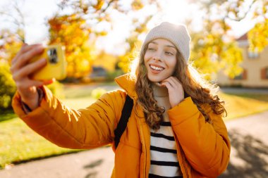 Parlak ceketli genç bir kadın güneşli bir günde telefonda selfie çekiyor. Güz parkından zevk alan ve telefonla yürüyen güzel bir kadın. Yürüyüş, eğlence konsepti.