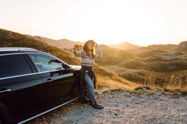 Telefonlu bir kadın blogcu gün batımında arabanın yanında canlı yayın yapıyor. Arabayla gezintiye çıkmış mutlu bir kadın ve güneşte telefon. Ulaşım ve macera konsepti.