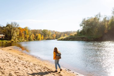 Parlak ceketli bir kadın gezgin güneşli bir günde nehir boyunca yürür. Sırt çantalı genç bir kadın dışarıda güneşli bir sonbahar gününün tadını çıkarıyor. Seyahat ve doğa kavramı.