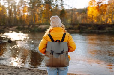 Parlak ceketli bir kadın gezgin güneşli bir günde nehir boyunca yürür. Sırt çantalı genç bir kadın dışarıda güneşli bir sonbahar gününün tadını çıkarıyor. Seyahat ve doğa kavramı.
