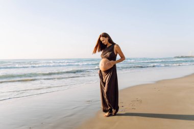 Güzel hamile kadın gün batımında deniz kıyısında duruyor. Deniz manzarasından hoşlanan, kıyıda yürüyen tatlı bir kadın. Yürüyüş, özgürlük ve zevk kavramı..