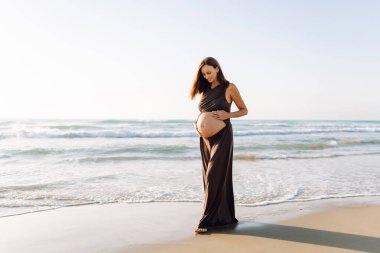 Güzel hamile kadın gün batımında deniz kıyısında duruyor. Deniz manzarasından hoşlanan, kıyıda yürüyen tatlı bir kadın. Yürüyüş, özgürlük ve zevk kavramı..