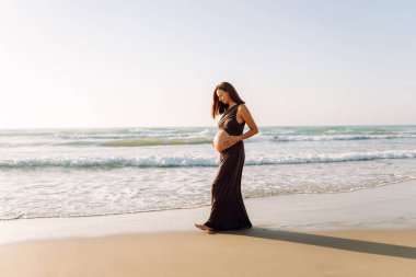 Güzel hamile kadın gün batımında deniz kıyısında duruyor. Deniz manzarasından hoşlanan, kıyıda yürüyen tatlı bir kadın. Yürüyüş, özgürlük ve zevk kavramı..