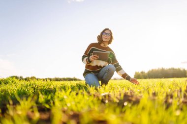 Dijital tabletli genç bir kadın çiftçi, yeşil tarım alanında güneşleniyor. Yeşil lahanalı dişi tarım uzmanı mahsulün büyümesini ve kalitesini kontrol ediyor. Çiftçilik ve teknoloji kavramı.