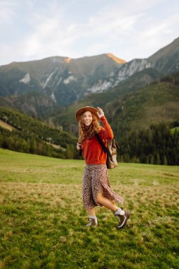 Sırt çantası ve şapkası olan bir bayan gezgin yeşil bir dağ manzarasının tadını çıkarıyor. Genç bir kadın tepenin üstünde yürüyor. Doğa ve macera kavramı, yürüyüş. Etkin yaşam biçimi.