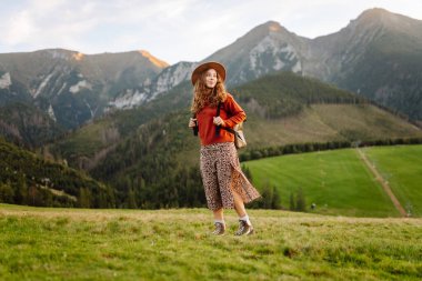 Sırt çantası ve şapkası olan bir bayan gezgin yeşil bir dağ manzarasının tadını çıkarıyor. Genç bir kadın tepenin üstünde yürüyor. Doğa ve macera kavramı, yürüyüş. Etkin yaşam biçimi.