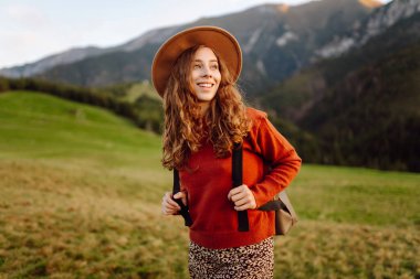 Sırt çantası ve şapkası olan bir bayan gezgin yeşil bir dağ manzarasının tadını çıkarıyor. Genç bir kadın tepenin üstünde yürüyor. Doğa ve macera kavramı, yürüyüş. Etkin yaşam biçimi.