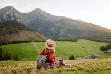 Sırt çantası ve şapkası olan bir bayan gezgin yeşil bir dağ manzarasının tadını çıkarıyor. Genç bir kadın tepenin üstünde yürüyor. Doğa ve macera kavramı, yürüyüş. Etkin yaşam biçimi.
