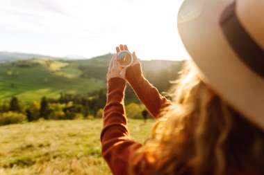 Güneşli bir günde sırt çantası ve pusula tutan kadın yürüyüşçünün arka görüntüsü. Yürüyüş yolu arayan kadın yürüyüşçü, gün batımının tadını çıkarıyor. Navigasyon kavramı.