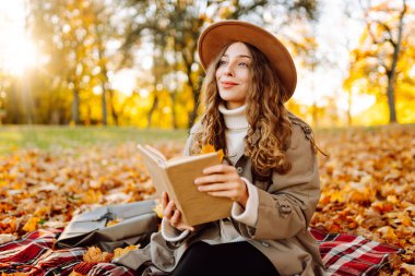 Şapkalı hoş bir kadın, sonbahar parkında yumuşak bir battaniyeye oturup kitap okur. Güzel bir kadın güneşli ormanda dinlenir, sarı yapraklarla çevrili bir çayırda oturur, doğanın tadını çıkarır. Dinlenme ve rahatlama kavramı.