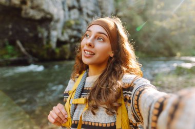 Sırt çantalı genç bayan yürüyüşçünün portresi gün batımında bir dağ nehrinin arka planında selfie çekiyor. Güneşli bir günde doğanın tadını çıkaran güzel bir gezgin. Teknoloji kavramı, blog yazma.
