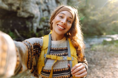 Sırt çantalı genç bayan yürüyüşçünün portresi gün batımında bir dağ nehrinin arka planında selfie çekiyor. Güneşli bir günde doğanın tadını çıkaran güzel bir gezgin. Teknoloji kavramı, blog yazma.