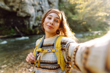 Sırt çantalı genç bayan yürüyüşçünün portresi gün batımında bir dağ nehrinin arka planında selfie çekiyor. Güneşli bir günde doğanın tadını çıkaran güzel bir gezgin. Teknoloji kavramı, blog yazma.