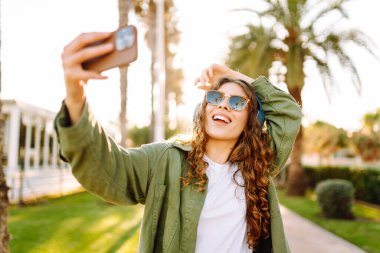 Telefonları ve kablosuz kulaklıkları olan neşeli bir kadın güneşli parkta selfie çekiyor. Güneş gözlüklü güzel kadın güneş ışınları altında yürüyüp blog yazmaktan zevk alıyor. Teknoloji ve keyif kavramı.
