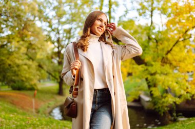 Şık bir trençkot giymiş neşeli bir kadın sonbahar parkında poz veriyor. Güzel bir kadın dışarıda yürümekten hoşlanır. Güzellik ve moda konsepti. Sonbahar havası.