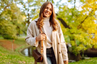 Şık bir trençkot giymiş neşeli bir kadın sonbahar parkında poz veriyor. Güzel bir kadın dışarıda yürümekten hoşlanır. Güzellik ve moda konsepti. Sonbahar havası.