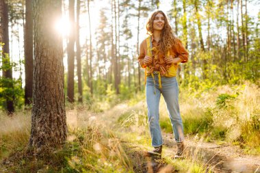 Parlak bir sırt çantası olan mutlu bir yürüyüşçü güneş ışınlarında bir ağacın altında oturur. Güzel bir kadın sonbahar ormanında bir maceranın tadını çıkarır. Kavram: yürüyüş, rahatlama, doğa. Etkin yaşam biçimi.