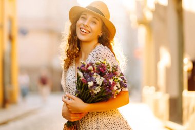 Bir buket kır çiçeğiyle şapkalı mutlu bir kadın güneşli bir şehir caddesinde geziniyor. Tatlı bir kadın gün batımını ve açık havada buketini sever. Güzellik ve yürüme kavramı.