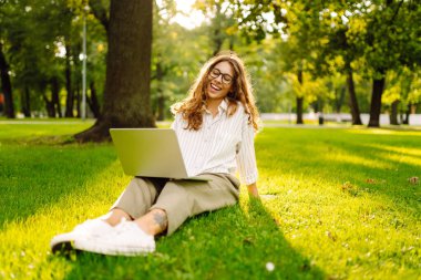 Gözlüklü, gülümseyen bir kadın elinde dizüstü bilgisayarla yeşil bir çayırda oturuyor. Dişi bir serbest çalışan dışarıda dizüstü bilgisayarıyla güneşli günün tadını çıkarıyor. Kavram: uzak çalışma, rahatlama.