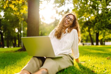 Gözlüklü, gülümseyen bir kadın elinde dizüstü bilgisayarla yeşil bir çayırda oturuyor. Dişi bir serbest çalışan dışarıda dizüstü bilgisayarıyla güneşli günün tadını çıkarıyor. Kavram: uzak çalışma, rahatlama.