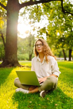 Gözlüklü, gülümseyen bir kadın elinde dizüstü bilgisayarla yeşil bir çayırda oturuyor. Dişi bir serbest çalışan dışarıda dizüstü bilgisayarıyla güneşli günün tadını çıkarıyor. Kavram: uzak çalışma, rahatlama.