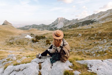 Sırt çantası ve şapkası olan genç bir bayan yürüyüşçü bir kayanın üzerinde oturur ve dağ manzarasına bakar. Yürüyüş parkurundaki bir kadın kendini özgür hisseder ve maceradan zevk alır. Seyahat kavramı, özgürlük..
