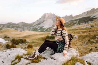 Sırt çantası ve şapkası olan genç bir bayan yürüyüşçü bir kayanın üzerinde oturur ve dağ manzarasına bakar. Yürüyüş parkurundaki bir kadın kendini özgür hisseder ve maceradan zevk alır. Seyahat kavramı, özgürlük..