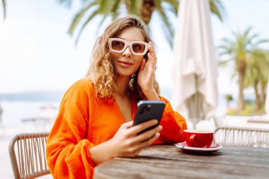 Telefonu olan güzel bir kadın yazlık terasta oturup palmiye ağaçlarının önünde kahve içiyor. Genç gezgin güneşte telefonuna mesaj atıyor. Güzellik, moda ve rahatlama kavramı.