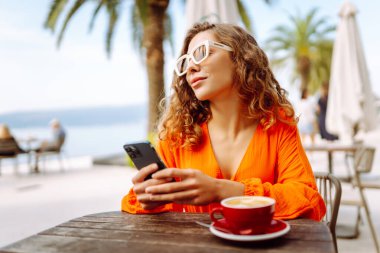 Telefonu olan güzel bir kadın yazlık terasta oturup palmiye ağaçlarının önünde kahve içiyor. Genç gezgin güneşte telefonuna mesaj atıyor. Güzellik, moda ve rahatlama kavramı.