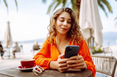 Telefonu olan güzel bir kadın yazlık terasta oturup palmiye ağaçlarının önünde kahve içiyor. Genç gezgin güneşte telefonuna mesaj atıyor. Güzellik, moda ve rahatlama kavramı.