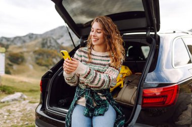 Şapkalı bir gezginin portresi elinde telefonla bir arabanın bagajında oturuyor. Neşeli bir kadın yolculukta dağ manzarasından zevk alır. Dağların, ulaşımın ve blog yazmanın kavramsal görüntüleri.