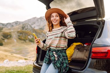 Şapkalı bir gezginin portresi elinde telefonla bir arabanın bagajında oturuyor. Neşeli bir kadın yolculukta dağ manzarasından zevk alır. Dağların, ulaşımın ve blog yazmanın kavramsal görüntüleri.