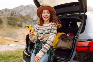 Şapkalı bir gezginin portresi elinde telefonla bir arabanın bagajında oturuyor. Neşeli bir kadın yolculukta dağ manzarasından zevk alır. Dağların, ulaşımın ve blog yazmanın kavramsal görüntüleri.