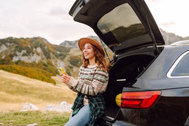 Şapkalı bir gezginin portresi elinde telefonla bir arabanın bagajında oturuyor. Neşeli bir kadın yolculukta dağ manzarasından zevk alır. Dağların, ulaşımın ve blog yazmanın kavramsal görüntüleri.