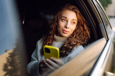 Neşeli bir kadın arabanın arka koltuğunda otururken telefonla konuşur. Telefonu olan genç bir kadın güneşli bir günde araba camından dışarı bakar. İletişim, teknoloji ve ulaşım kavramları.