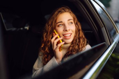 Neşeli bir kadın arabanın arka koltuğunda otururken telefonla konuşur. Telefonu olan genç bir kadın güneşli bir günde araba camından dışarı bakar. İletişim, teknoloji ve ulaşım kavramları.