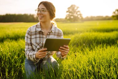 Dijital tabletli genç bir kadın. Güneş ışınlarında yeşil tarımsal bir alanda. Dişi bir çiftçi ekinlerin büyümesini tabletle kontrol eder ve günbatımının tadını çıkarır. Teknoloji kavramı, tarım..