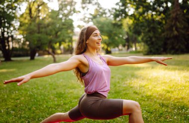 Atletik bir kadın güneşli bir parkta minder üzerinde yoga yapıyor. Spor kıyafetleri içinde güzel bir kadın egzersiz yapıyor, güneş ışınlarında egzersiz yapıyor. Konseptler: fitness, doğa ve vücut bakımı.